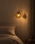 Riva amber blown-glass wall sconce on a matte black arm beside a bed, glowing without glare